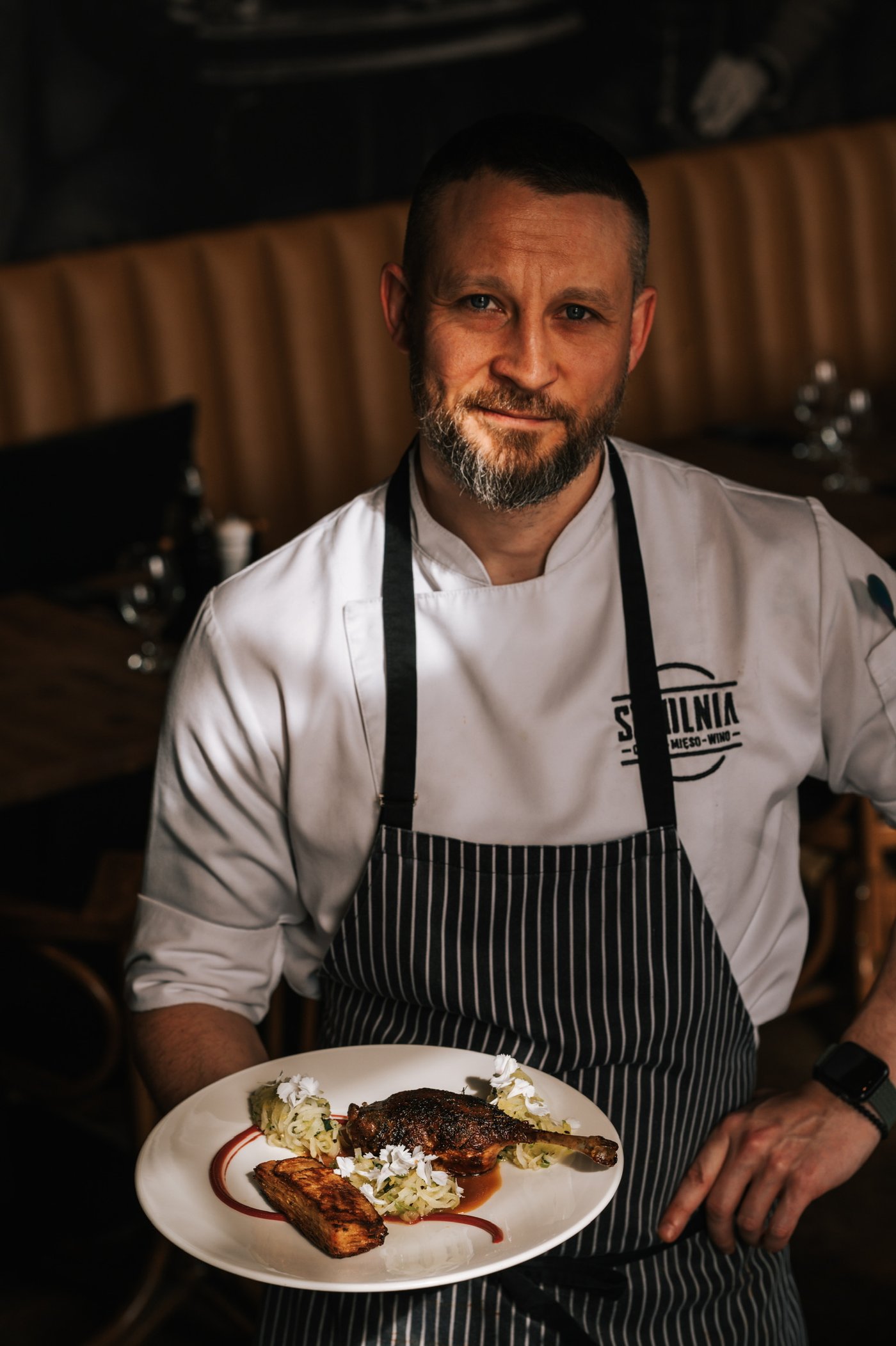 Dawid Golec, szef kuchni Sztolni — z talerzem konfitowanej kaczki we wnętrzu restauracji, pasiasty fartuch i biała kurtka z logo Sztolnia